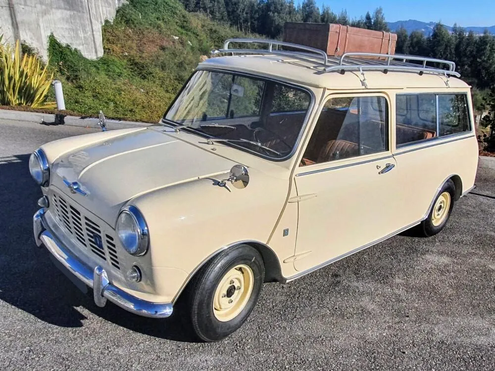 A cream-coloured vintage Morris Mini estate car with a roof rack and leather suitcase, parked on a hillside road